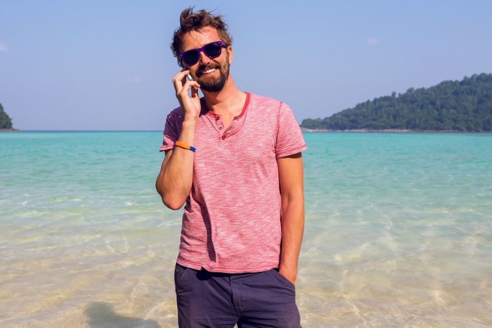 Hombre en la playa con teléfono en la mano y sonriendo tras un buen cuidado capilar